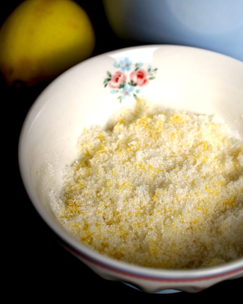 Lemon zest rubbed into sugar in a small floral bowl, with whole lemon and blue mixing bowl behind