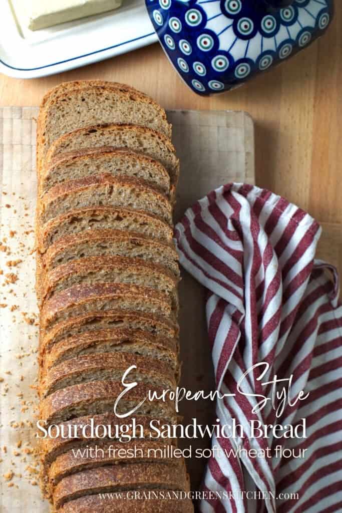 Top-down view of a whole European-style sourdough sandwich bread loaf made with fresh milled whole wheat flour, sliced and fanned on a cutting board, with tea towel and butter dish nearby.