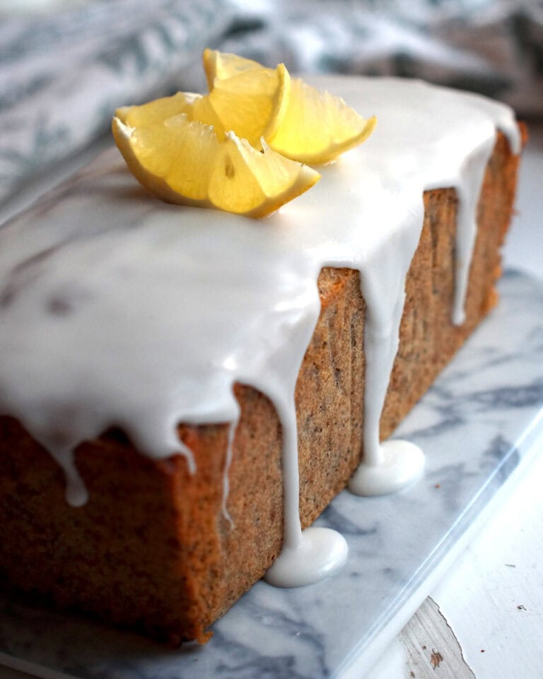 Glazed lemon poppy seed loaf on a marbled board, set on chipped white wood with a tea towel in the background