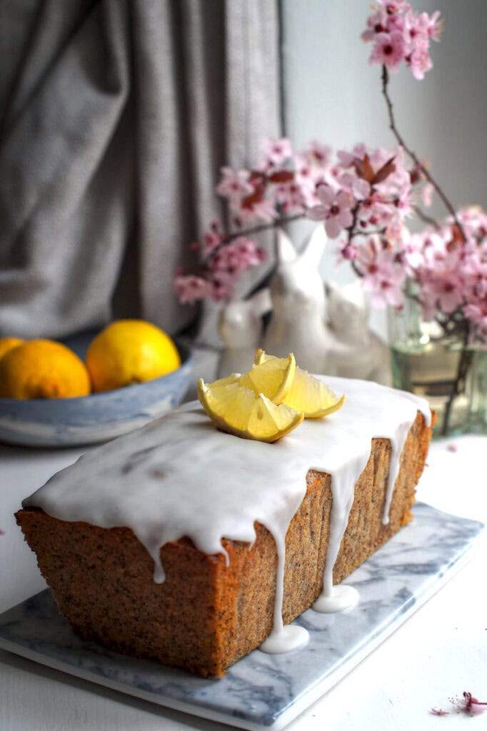Side view of lemon poppy seed loaf made with fresh milled flour, white glaze dripping, decorated with lemon slices, cherry blossoms, ceramic bunnies, and a bowl of lemons in the background