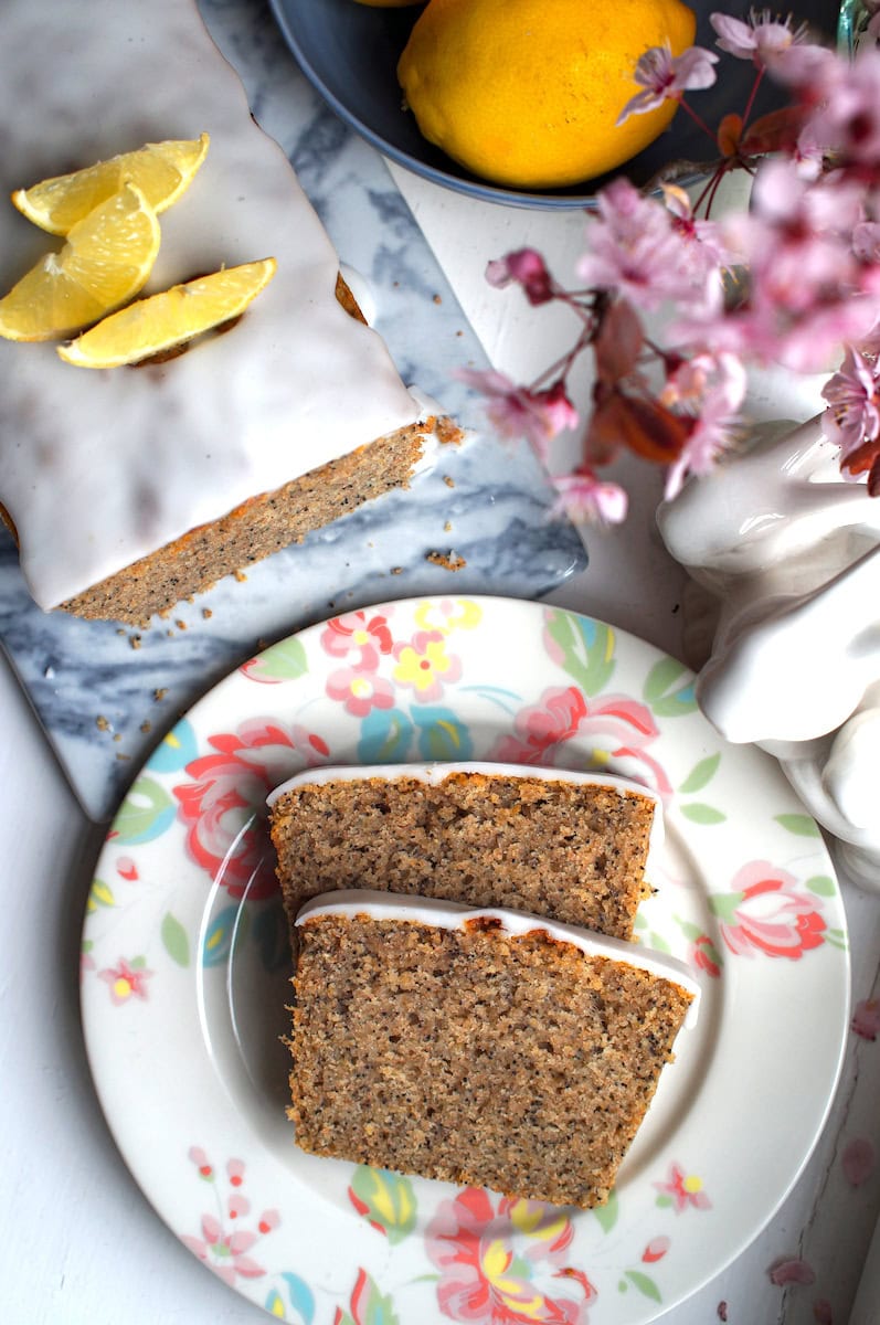 Overhead view of lemon poppy seed loaf made with fresh milled flour on a marbled board, two slices on a floral plate, bronzed crumb and white glaze, decorated for Easter with ceramic bunnies, lemons, and cherry blossoms at edges