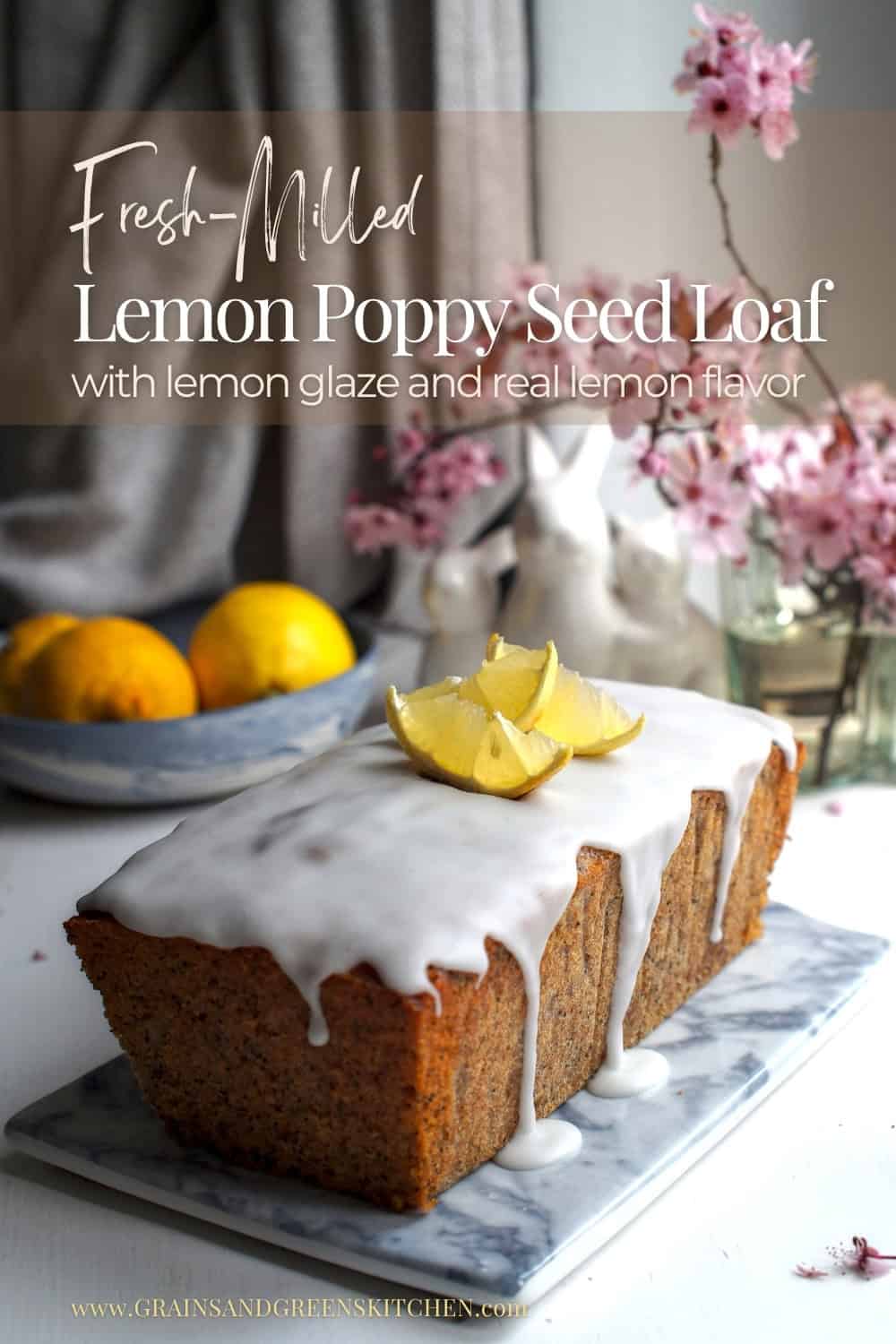 Side view of lemon poppy seed loaf made with fresh milled flour, white glaze dripping, decorated with lemon slices, cherry blossoms, ceramic bunnies, and a bowl of lemons in the background