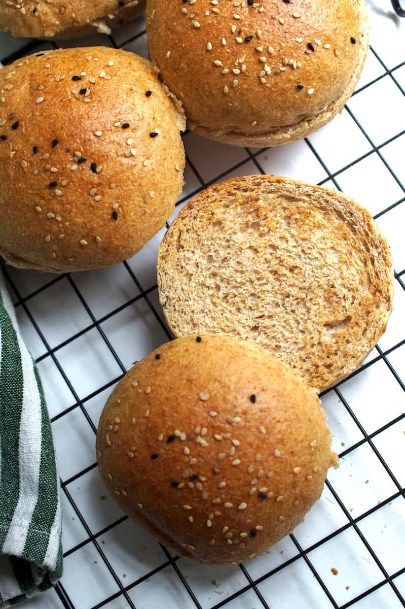 Freshly milled sourdough hamburger buns cooling on a rack, with a sliced and toasted bun showing the soft, even crumb inside.