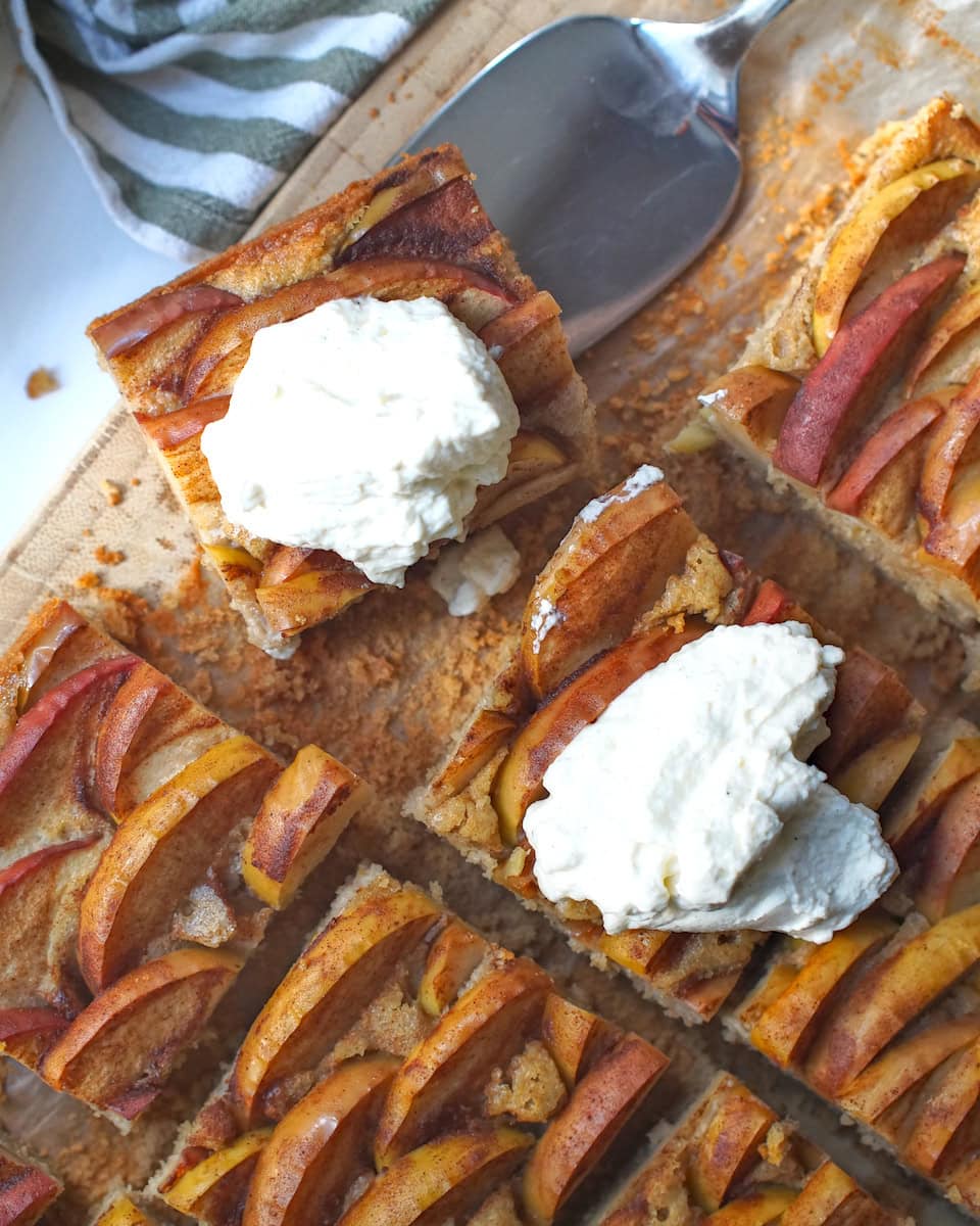 Overhead view of golden sourdough apple cake cut into squares, and two pieces topped with a dollop of bright white whipped cream.
