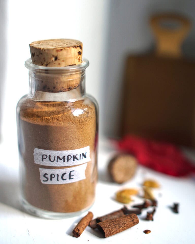 Homemade pumpkin pie spice blend in a clear glass jar with cork, surrounded by whole spices and fall leaves on a white surface