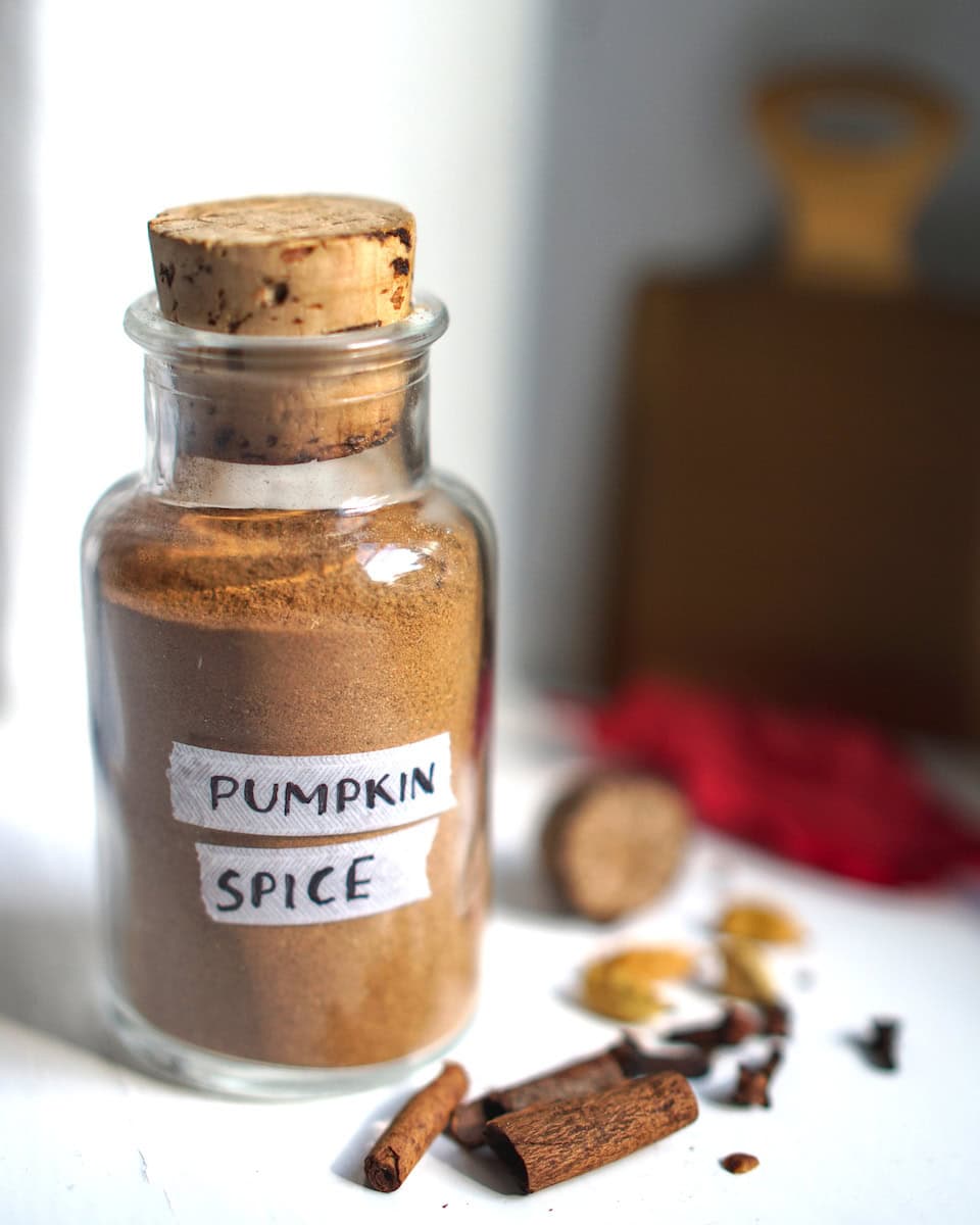 Homemade pumpkin pie spice blend in a clear glass jar with cork, surrounded by whole spices and fall leaves on a white surface