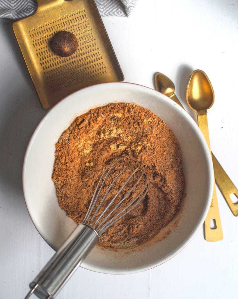 Top-down view of brown and yellow spices being mixed in a white bowl, with a nutmeg and golden grater and measuring spoons beside it